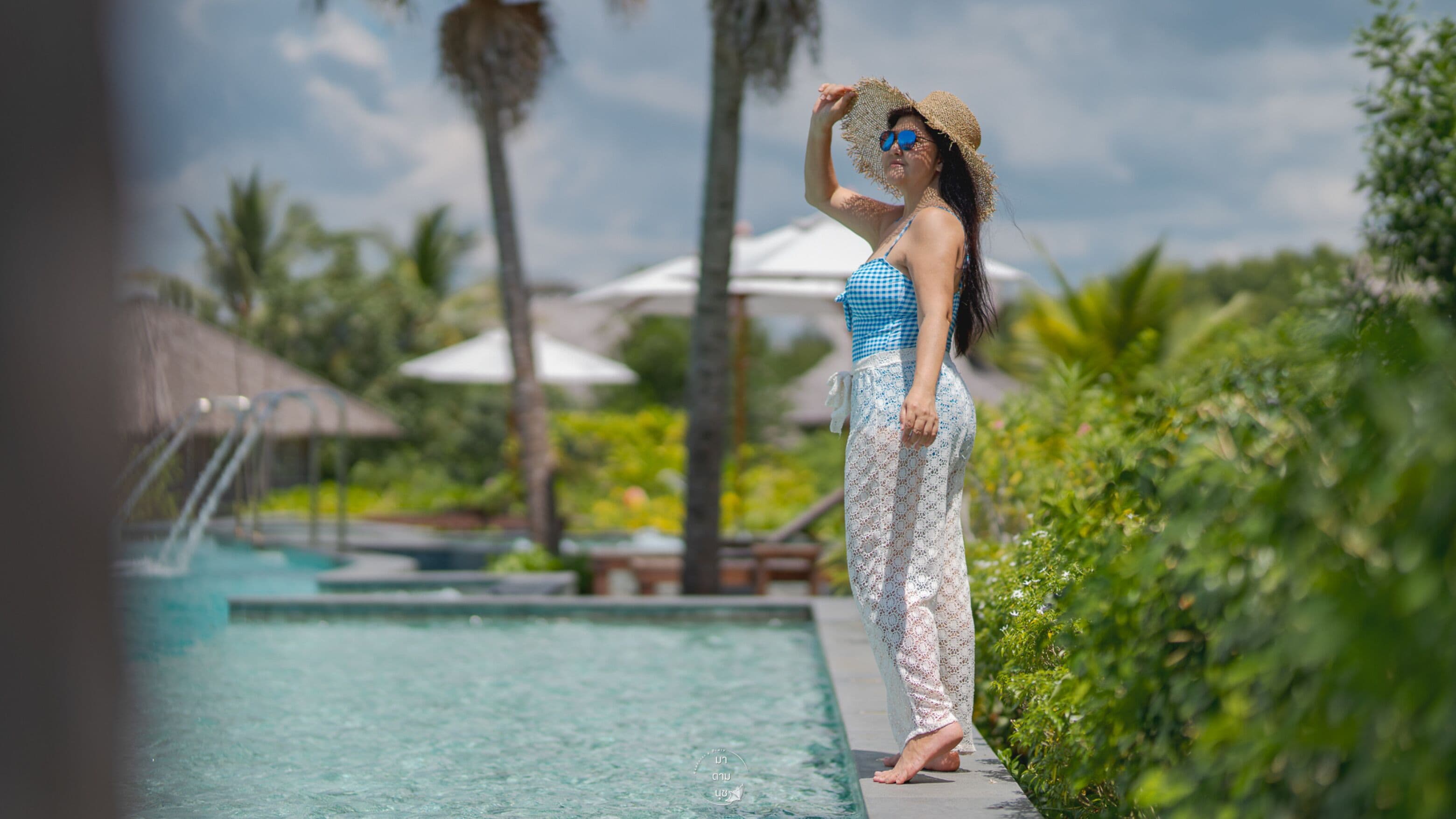 Eine Person steht an einem Pool in tropischer Umgebung, mit Palmen und Sonnenschirmen.