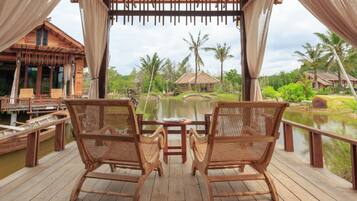 A wooden deck with two wicker chairs and a small table, overlooking a pond with palm trees and a thatched-roof hut in the background.