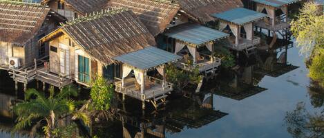 A cluster of traditional wooden houses with thatched roofs, built on stilts over water, surrounded by lush greenery.