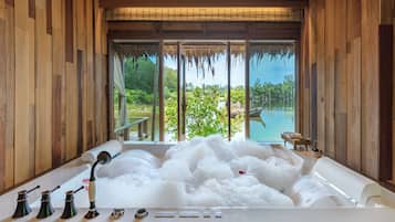 Een bad vol bubbels, een raam met uitzicht op een bos en water, en een houten interieur.