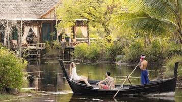 A boat with two people, one seated and one standing, on a calm river surrounded by lush greenery and wooden structures.