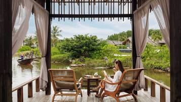 A wooden deck with two wicker chairs and a small table, overlooking a calm water body with boats and greenery.