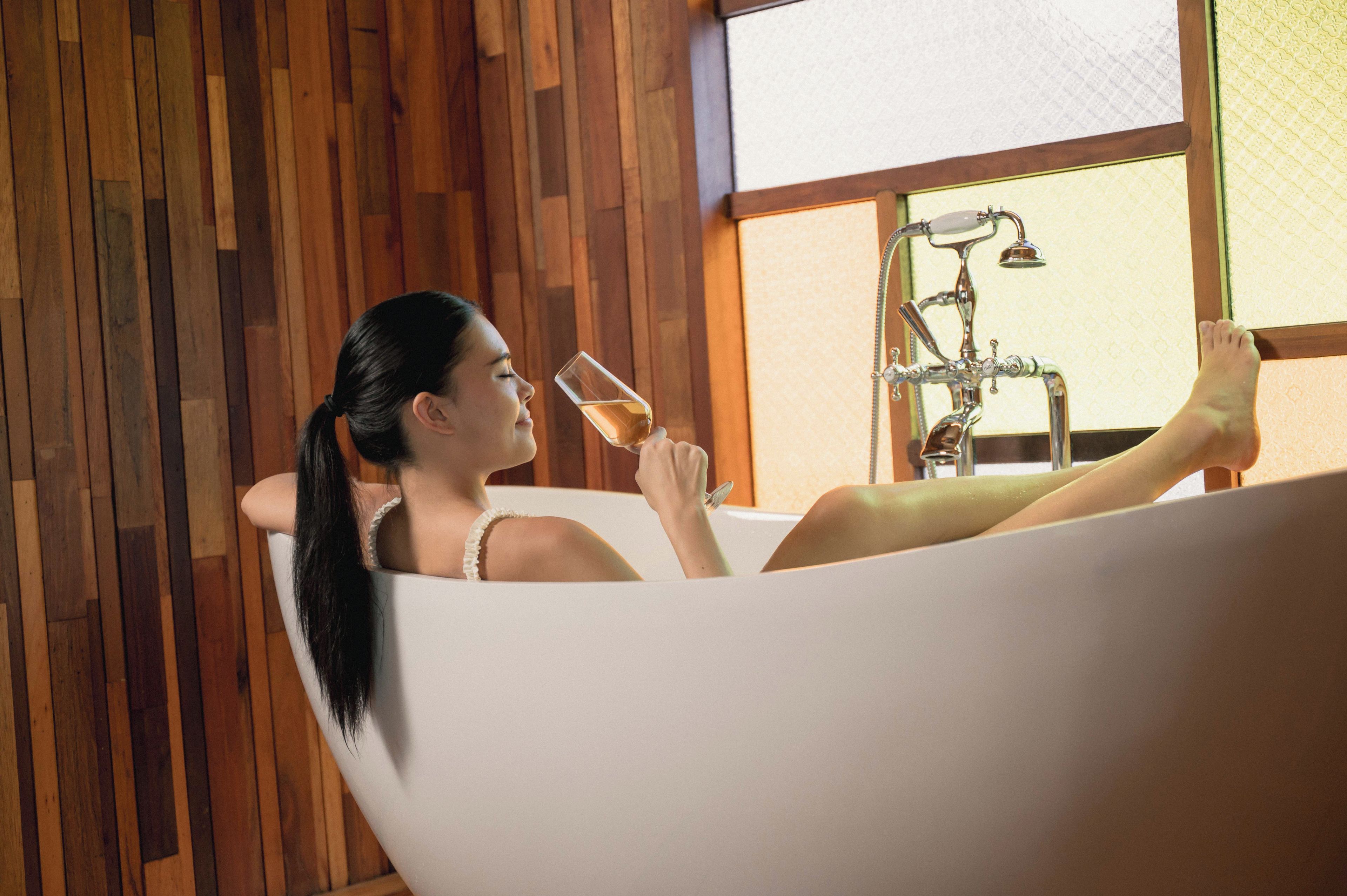 A person relaxing in a bathtub with a window and a shower in the background.
