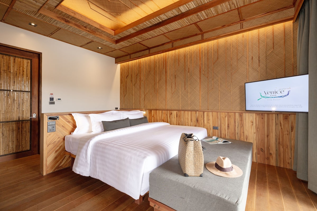 A hotel room with a bed, a grey sofa, a hat, and a television displaying 'Venice'.