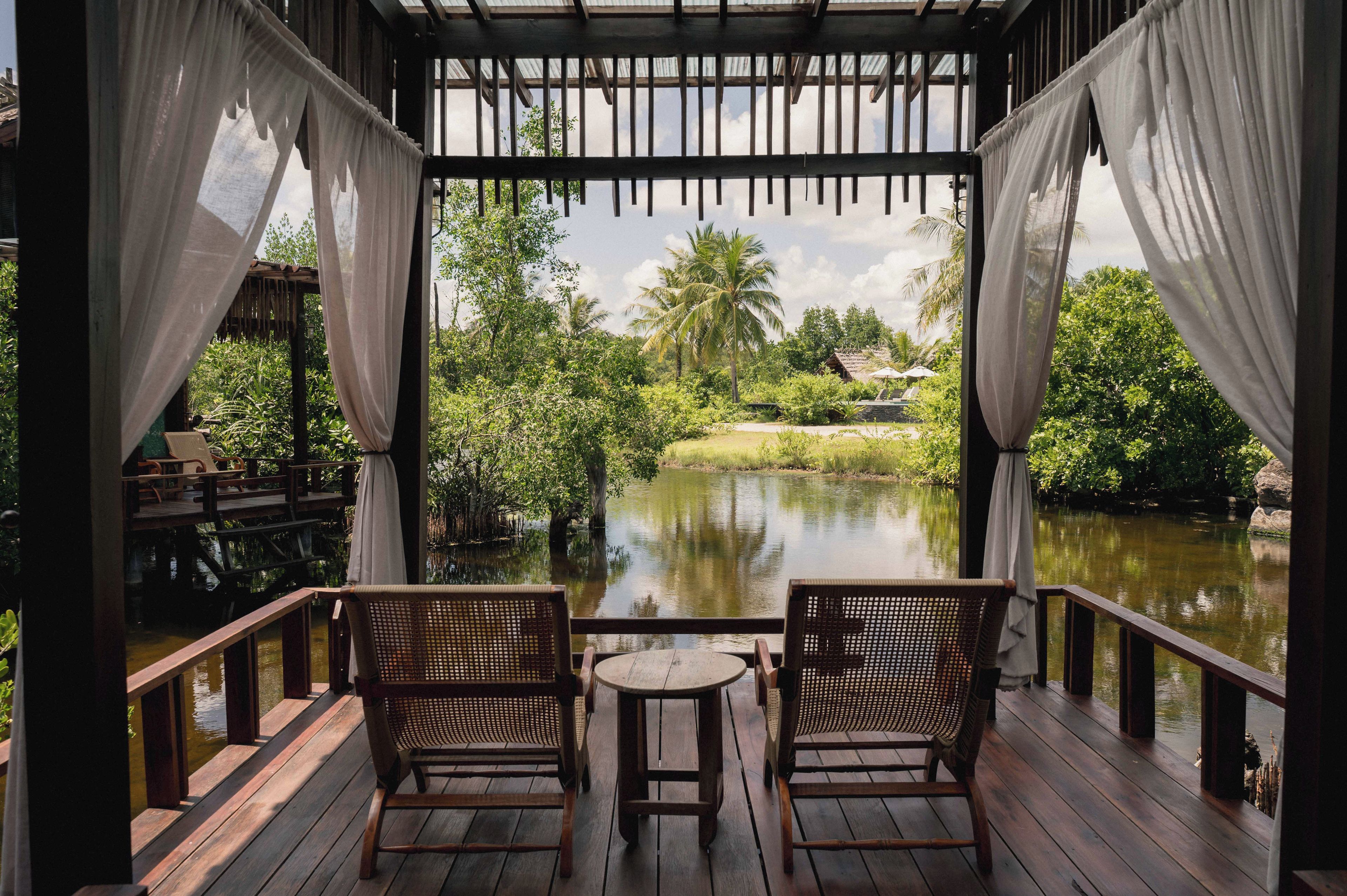 A wooden deck with two wicker chairs and a small table, overlooking a calm water body with lush greenery and a clear sky.