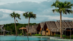 Un resort con piscina, cabañas de techo de paja, palmeras y un telón de fondo boscoso.