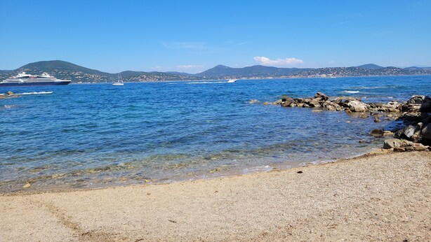 A coastal scene with a cruise ship, rocky shoreline, and distant hills.