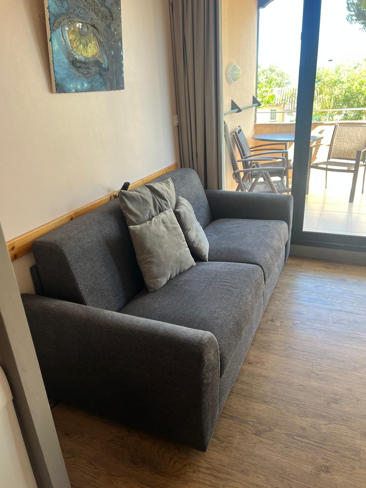 Een moderne woonkamer met een grijze bank, een houten vloer en een groot raam met uitzicht op tuinmeubilair.