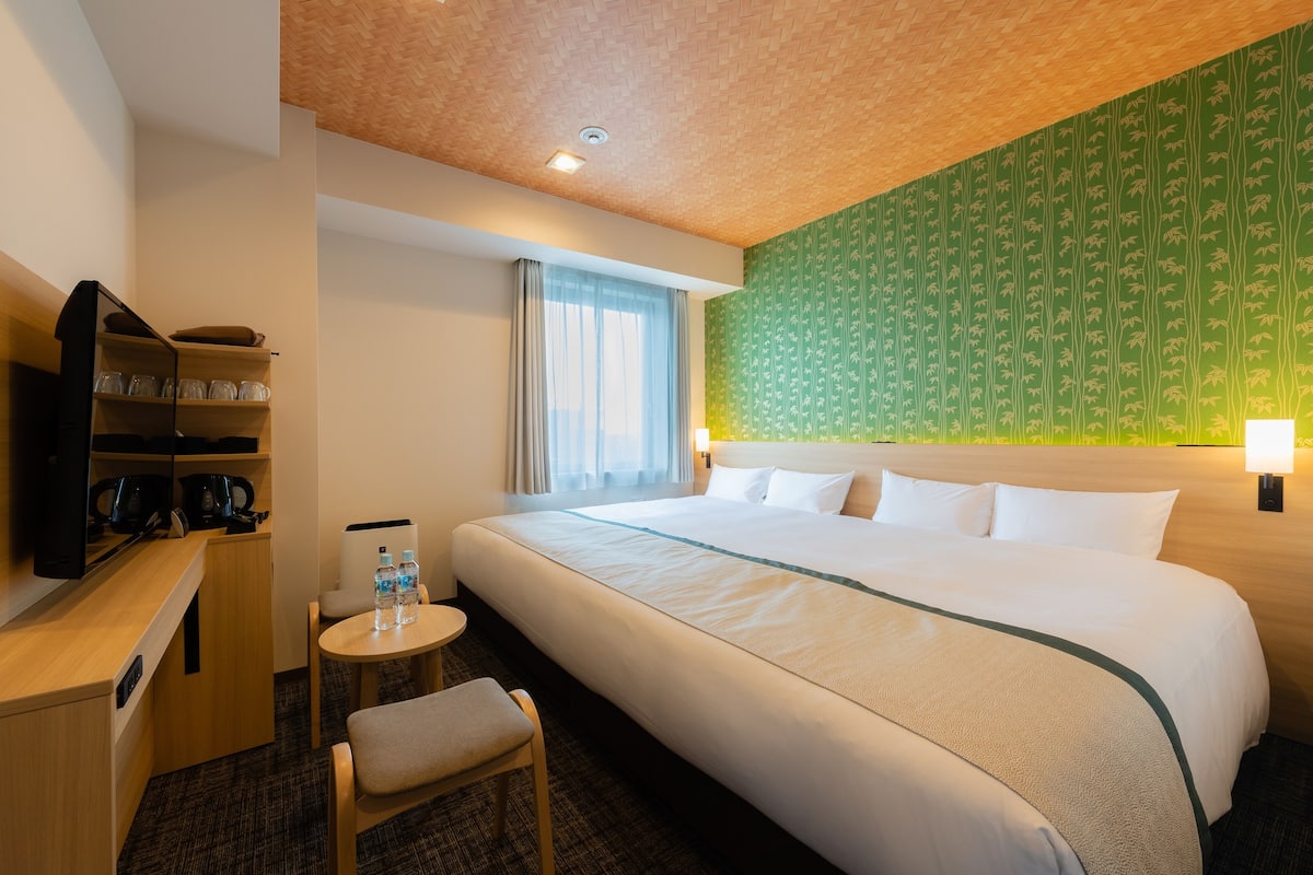 A hotel room with a large bed, a desk with a chair, a small table with a chair, a TV, and a shelf with glasses.