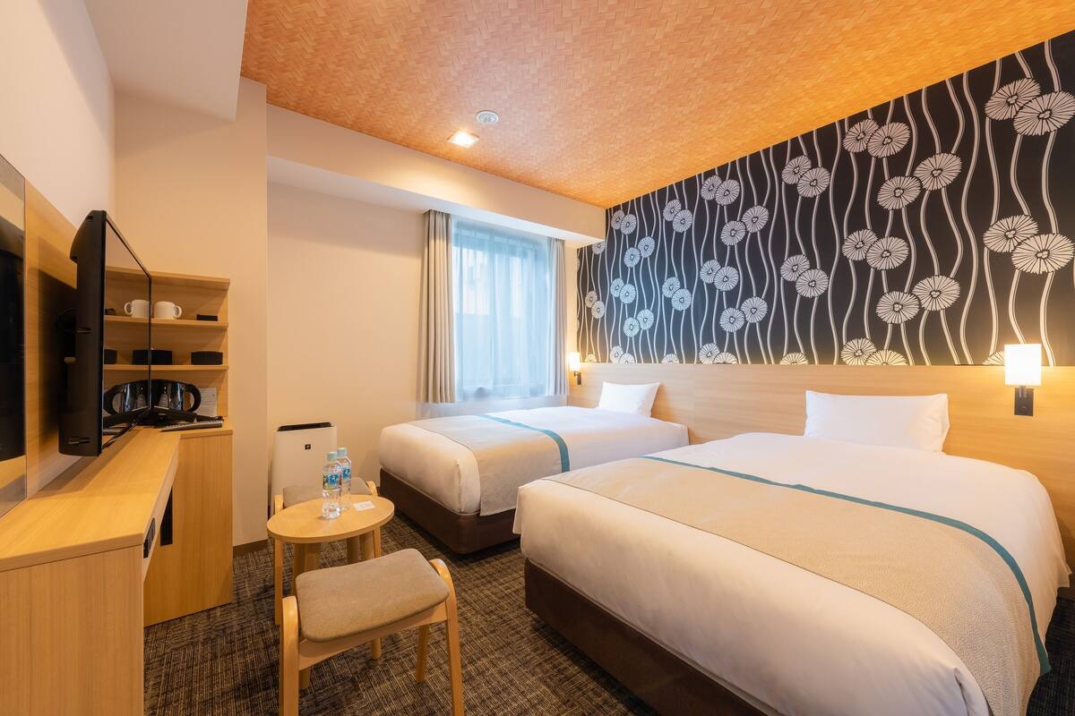 A hotel room with two beds, a desk, a chair, a TV, and a wall with a floral pattern.