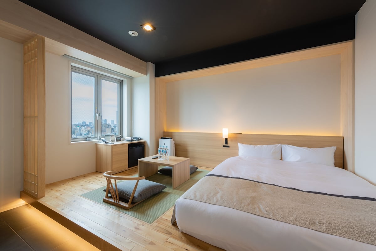 A hotel room with a large bed, a desk with a chair, a small table, a nightstand, and a window with a city view.