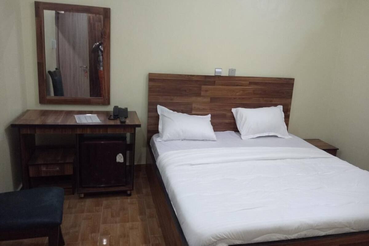 A bedroom with a wooden headboard, a bed with white bedding, a desk with a chair, and a small cabinet.