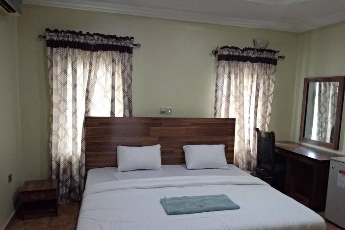 A bedroom with a wooden headboard, two white pillows, a bed with a blue towel, a desk with a chair, and curtains with a geometric pattern.
