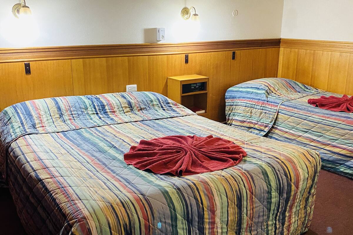 A hotel room with two beds, each with striped bedding and a red towel on the center. There is a wooden headboard, a nightstand with a radio, and a wall-mounted thermostat.