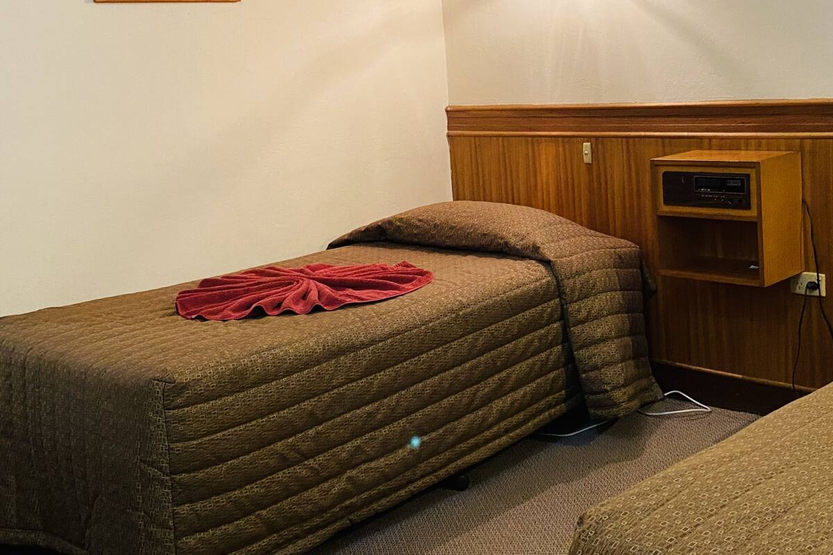 A single bed with a quilt and a red blanket, a wooden cabinet with a radio, and a framed picture on the wall.