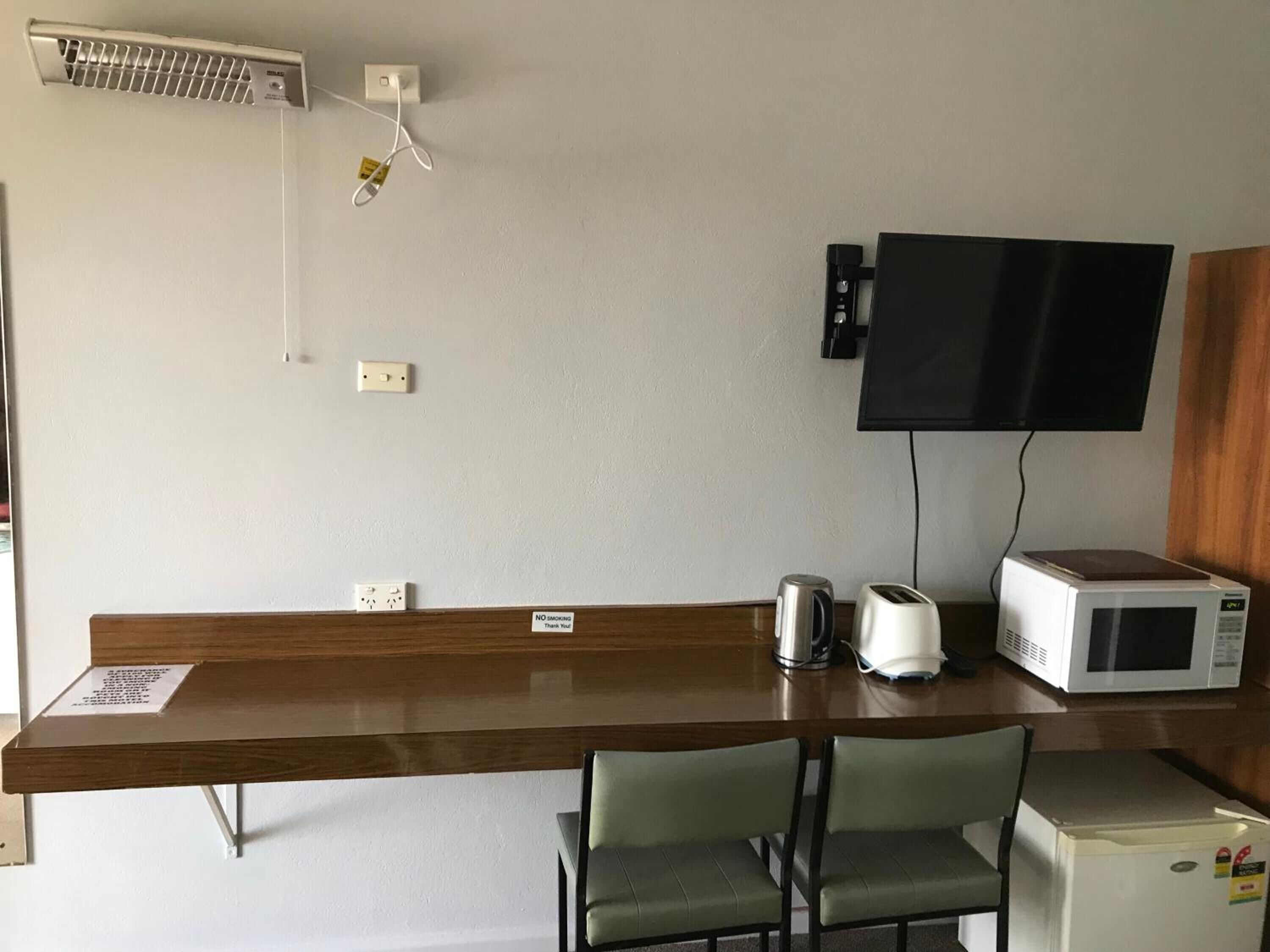 A hotel room with a wooden desk, two green chairs, a microwave, a kettle, and a television mounted on the wall.