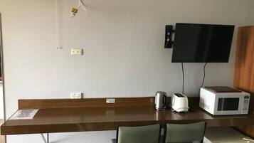 A hotel room with a wooden desk, two green chairs, a microwave, a kettle, and a television mounted on the wall.