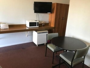 A small kitchenette with a microwave, toaster, and refrigerator. There is a flat-screen TV mounted on the wall above the counter. A round table with two chairs is positioned in front of the counter.