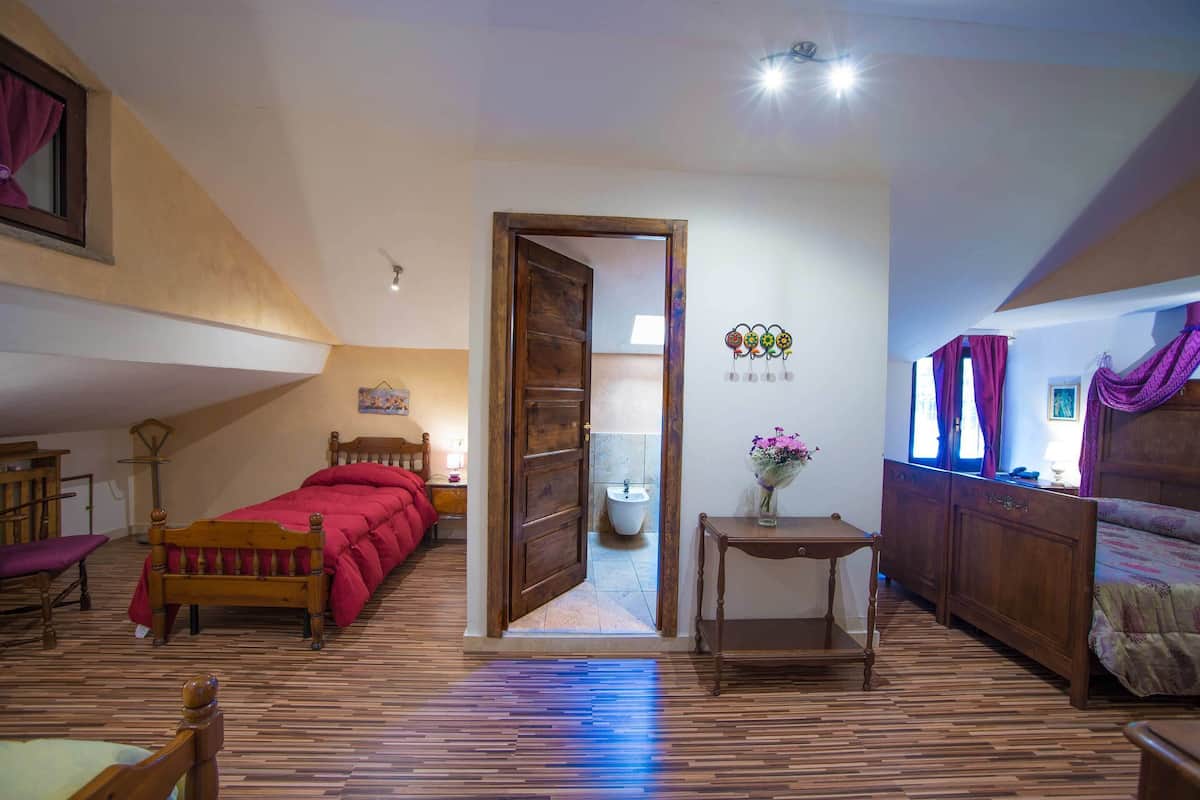 Une chambre avec un plancher en bois, un lit recouvert d’une couverture rouge, une armoire en bois, une petite table avec un vase, et une porte donnant sur une salle de bain.