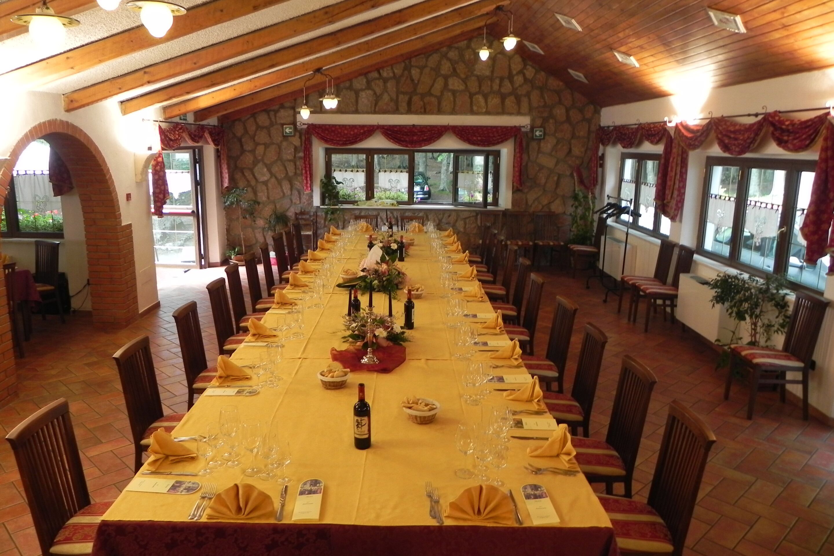 Una larga mesa de comedor dispuesta para una comida, con sillas alrededor, en una habitación con paredes de piedra y vigas de madera.