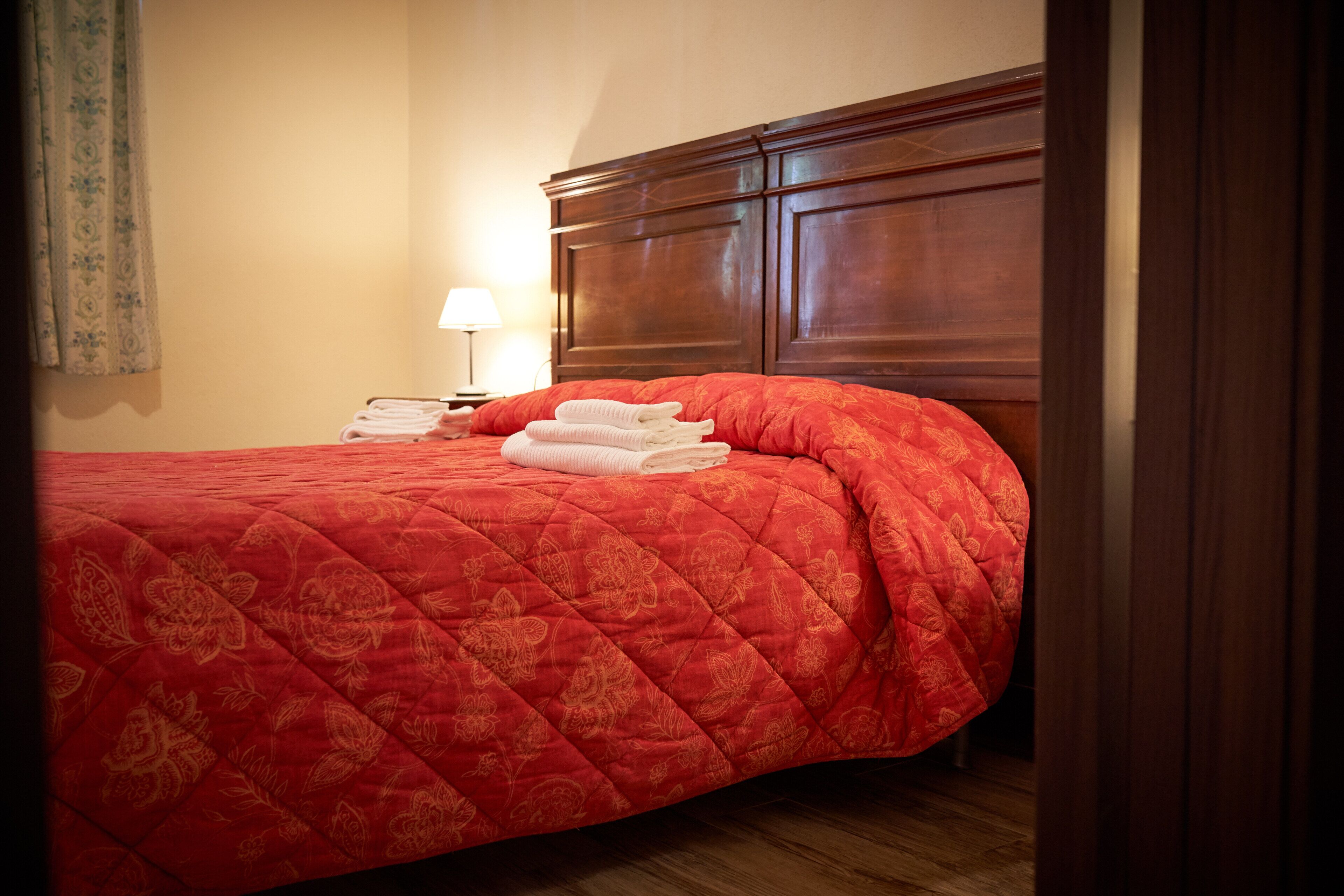 Ein ordentlich bezogenes Bett mit roter Steppdecke, einem Holz-Kopfteil und einem Nachttisch mit Lampe und Handtüchern.