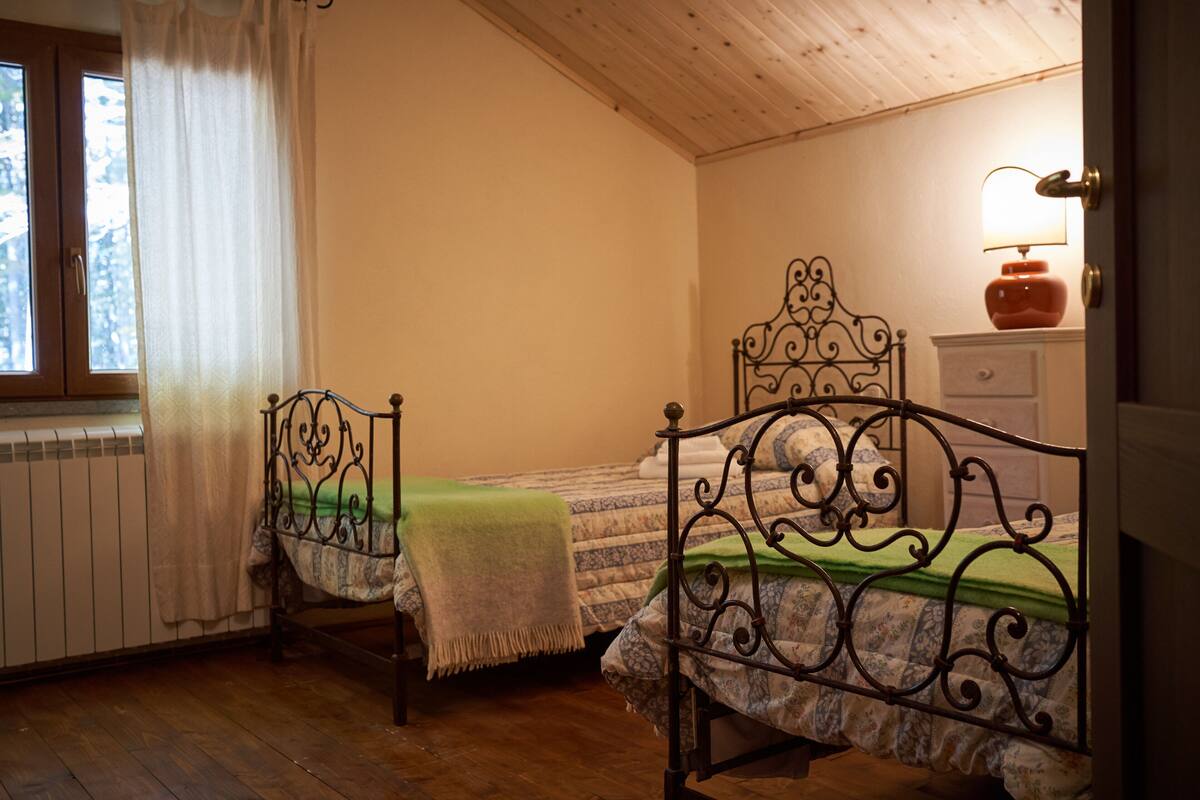 A bedroom with two beds, a radiator, a window with curtains, and a lamp on a bedside table.