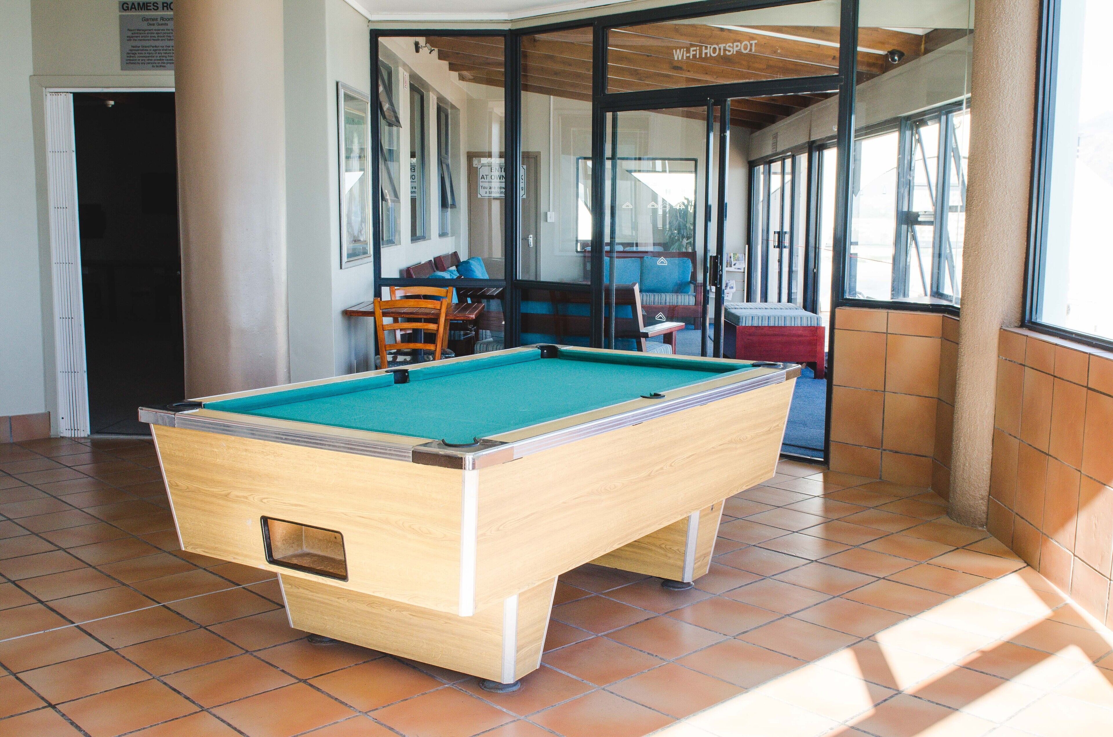 Une table de billard dans une pièce dont le sol est carrelé, avec un banc et une porte portant l’inscription « Salle de jeux ».