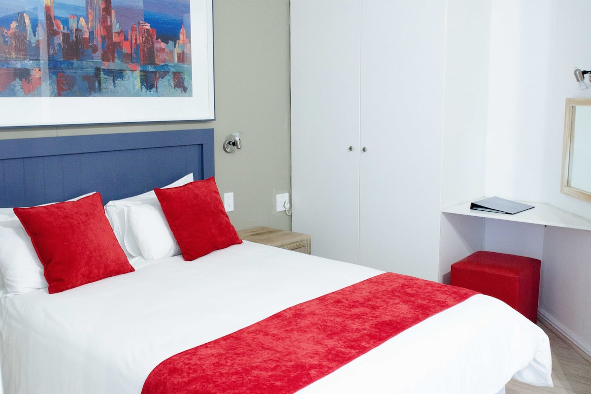 A hotel room with a bed, red pillows, a red throw, a white nightstand, a red stool, a white wardrobe, and a cityscape painting on the wall.