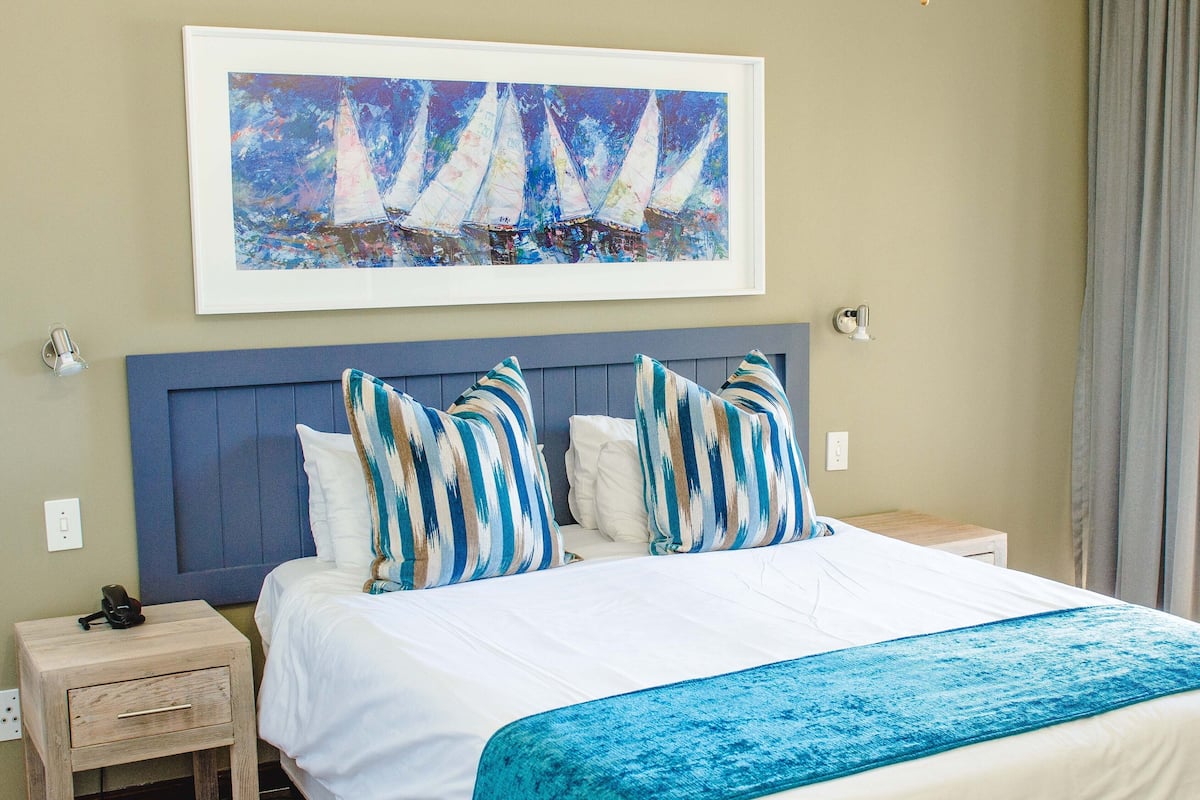 A neatly made bed with striped pillows, a framed painting of sailboats above it, and a bedside table with a lamp.