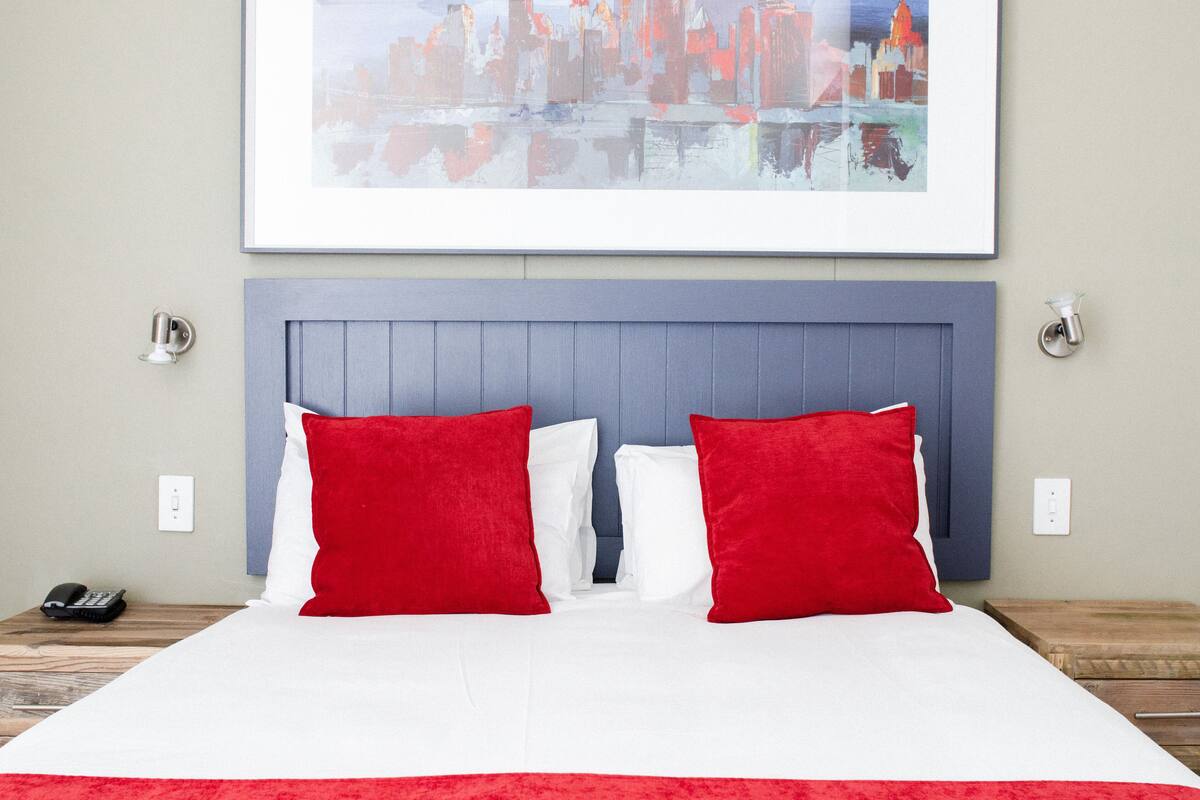 A neatly made bed with red and white pillows, a cityscape painting above, and a wooden nightstand with a phone.