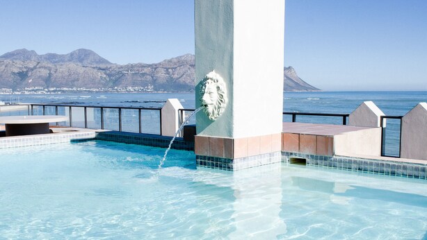 A swimming pool with clear blue water, a stone wall with a lion head sculpture, and a view of the ocean and mountains in the background.