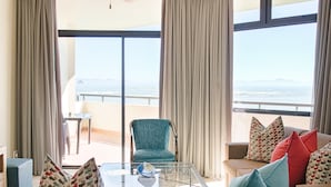 A living room with a sofa, armchairs, and a glass coffee table. There is a view of the ocean through large windows.