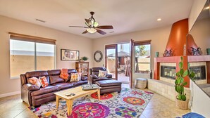 A living room with a brown leather sofa, a wooden coffee table, and a colorful rug. There is a ceiling fan, a fireplace, and a large window. - 222396 3BR Escape w/ Fire Pit: 1 Mi to Old Town (La Quinta)