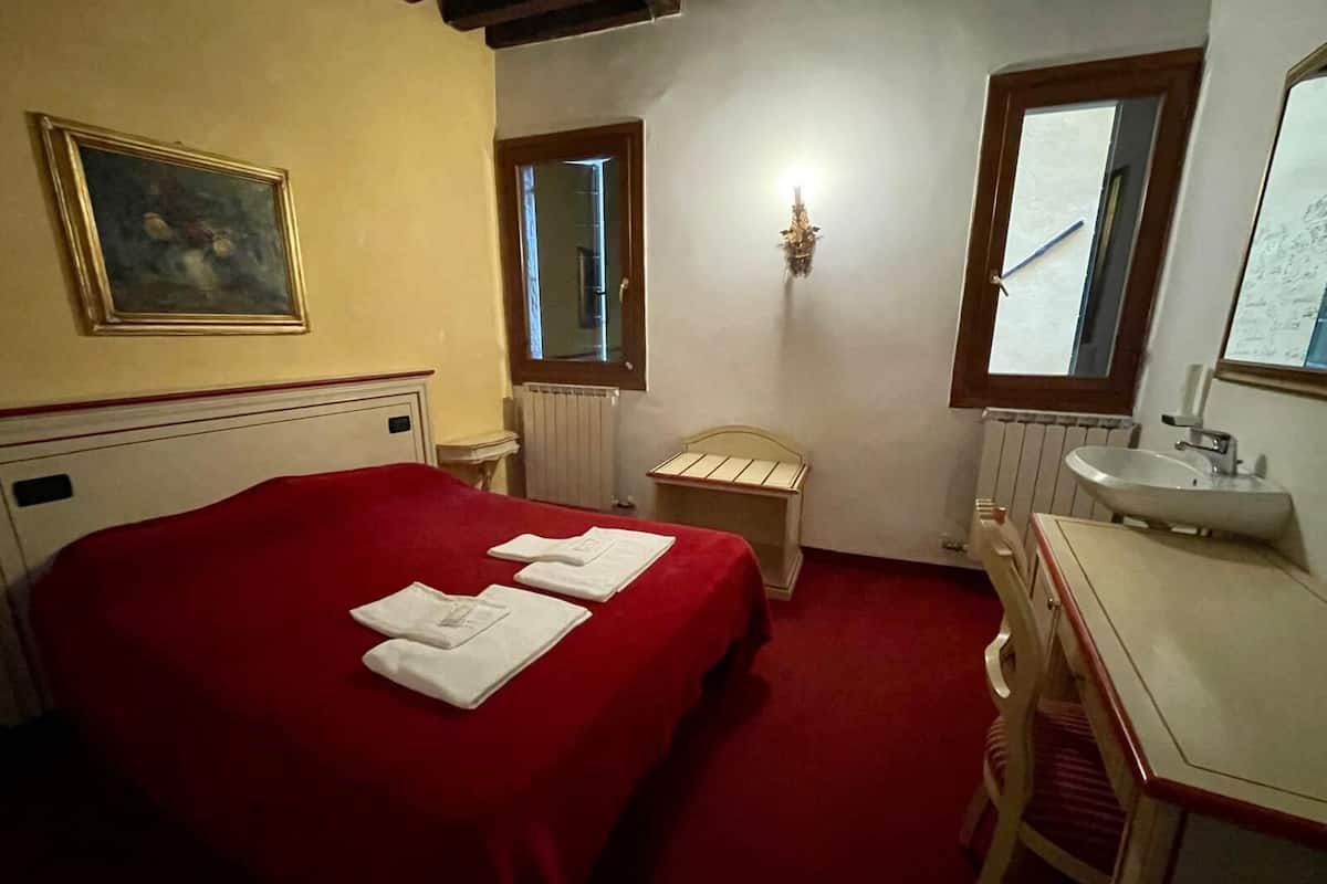 A hotel room with a bed, a bedside table, a small bedside table with a lamp, a window, a mirror, and a bathroom with a sink.