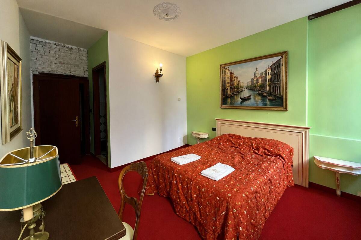 A hotel room with a bed, a desk, a chair, a lamp, a painting, and a door.