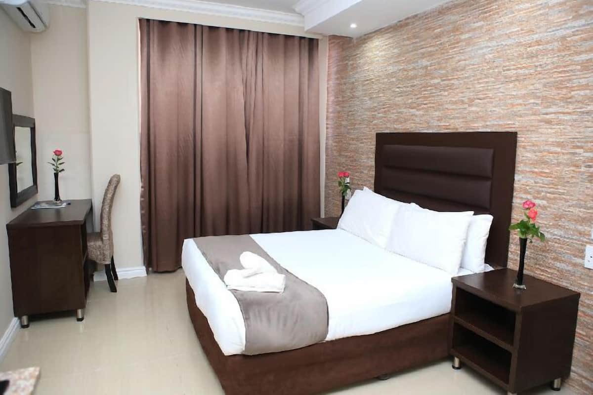 Een hotelkamer met een groot bed, een houten hoofdbord, een bruin gordijn, een klein bureau met een bloevas en een muur met een steenstructuur.