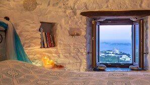 A cozy room with a view of a coastal town, featuring a bed, a small shelf with books, and a lit candle. - Anemomylos-Windmill (Syros)