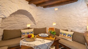 A cozy room with a stone wall, a wooden bench, and a cushioned sofa. There is a tray with fruits and a bowl on the bench, and a small table with a lace tablecloth. - Anemomylos-Windmill (Syros)