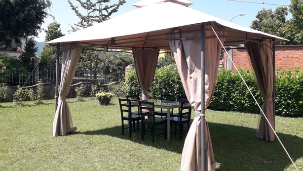 A covered outdoor seating area with a table and chairs, surrounded by greenery and a brick wall. - B&B Le Mi' Cocche (Capannori)
