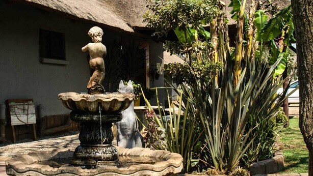 Una fuente de piedra con una estatua, rodeada de plantas tropicales y un edificio con techo de paja.