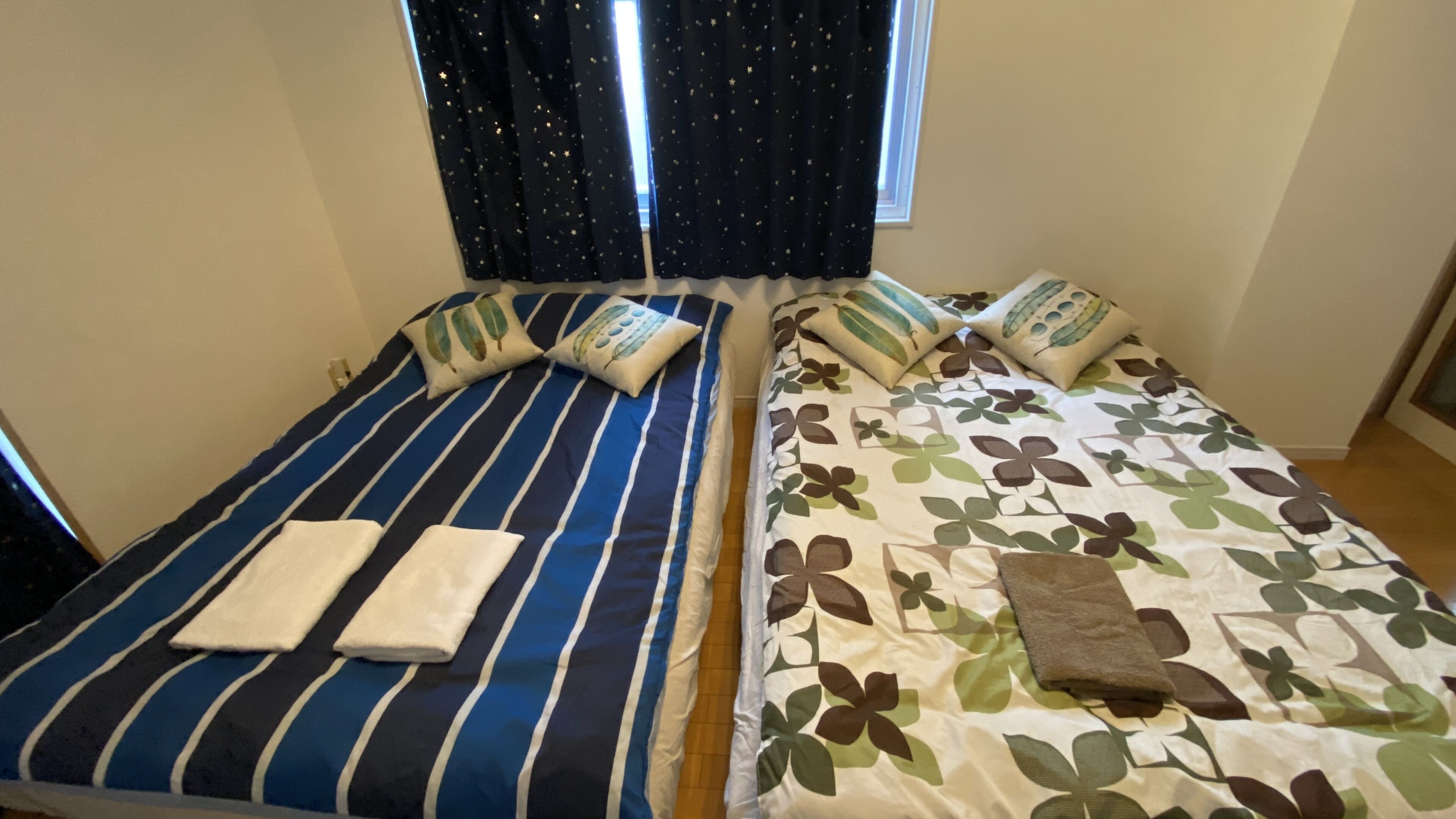 Two single beds with striped and floral bedding, a window with star-patterned curtains, and a wooden floor.