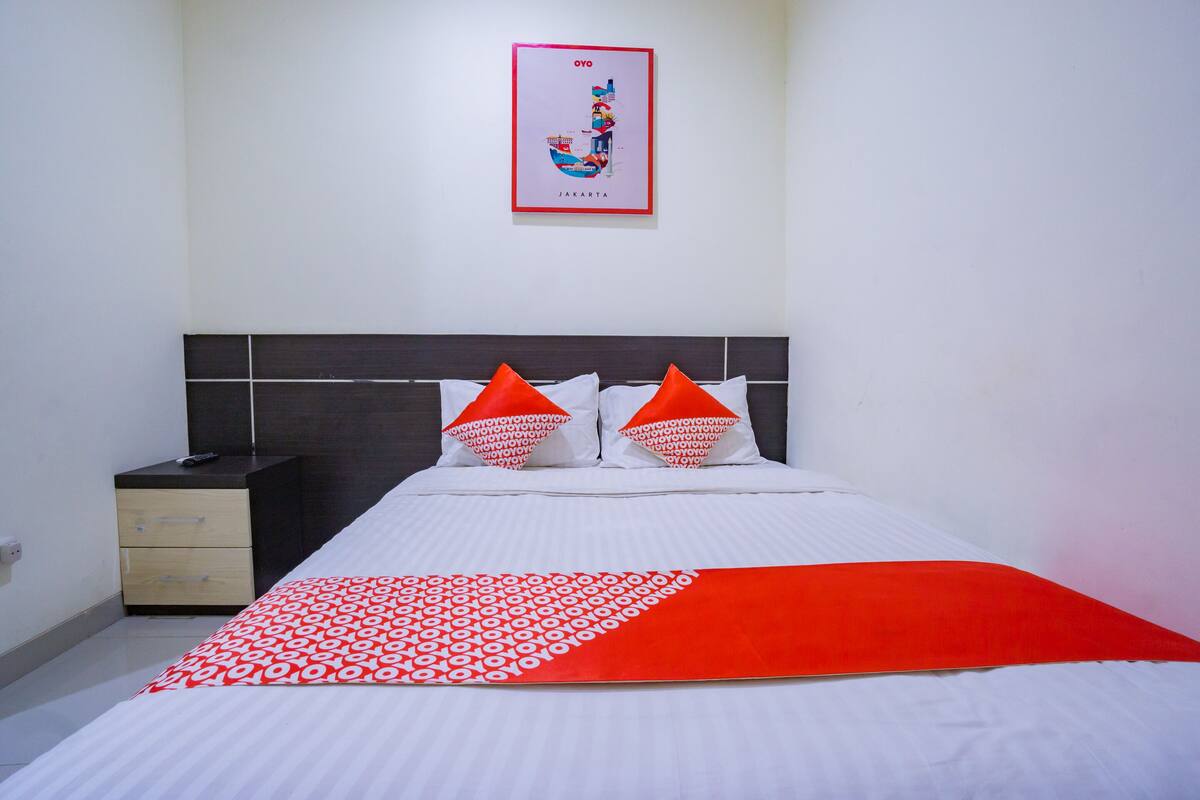 A neatly made bed with a white and red bedspread, two orange pillows, and a framed map of Jakarta on the wall.