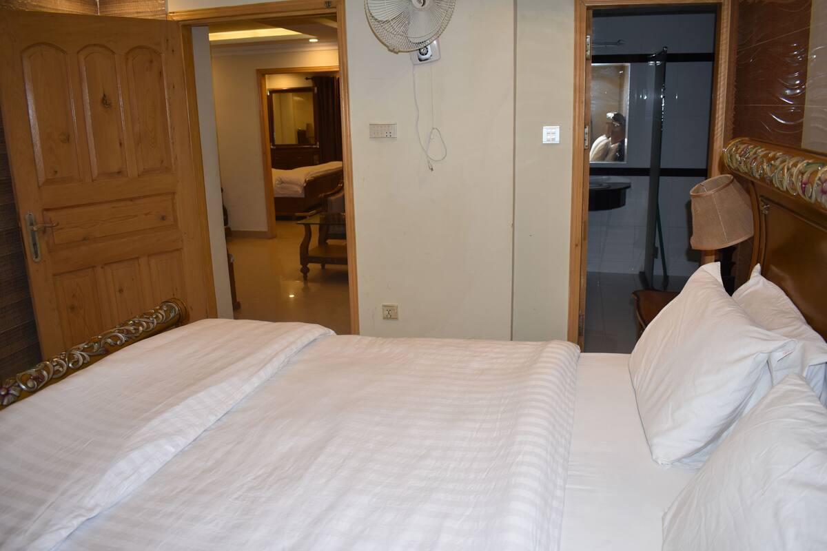 A hotel room with a wooden door, a bed with white bedding, a standing fan, and a mirror on the wall.