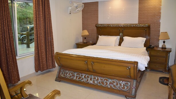 A bedroom with a wooden bed, two bedside tables with lamps, a large window with curtains, and a view of a parked car outside. - Hotel Faran Murree (Murree)