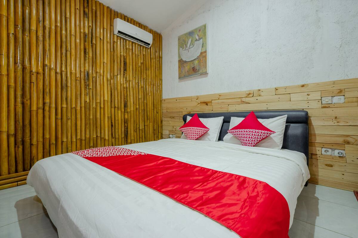 Un lit bien fait, recouvert d’une couvre-lit rouge et blanche, avec une tête de lit en bois et un panneau mural en bambou.