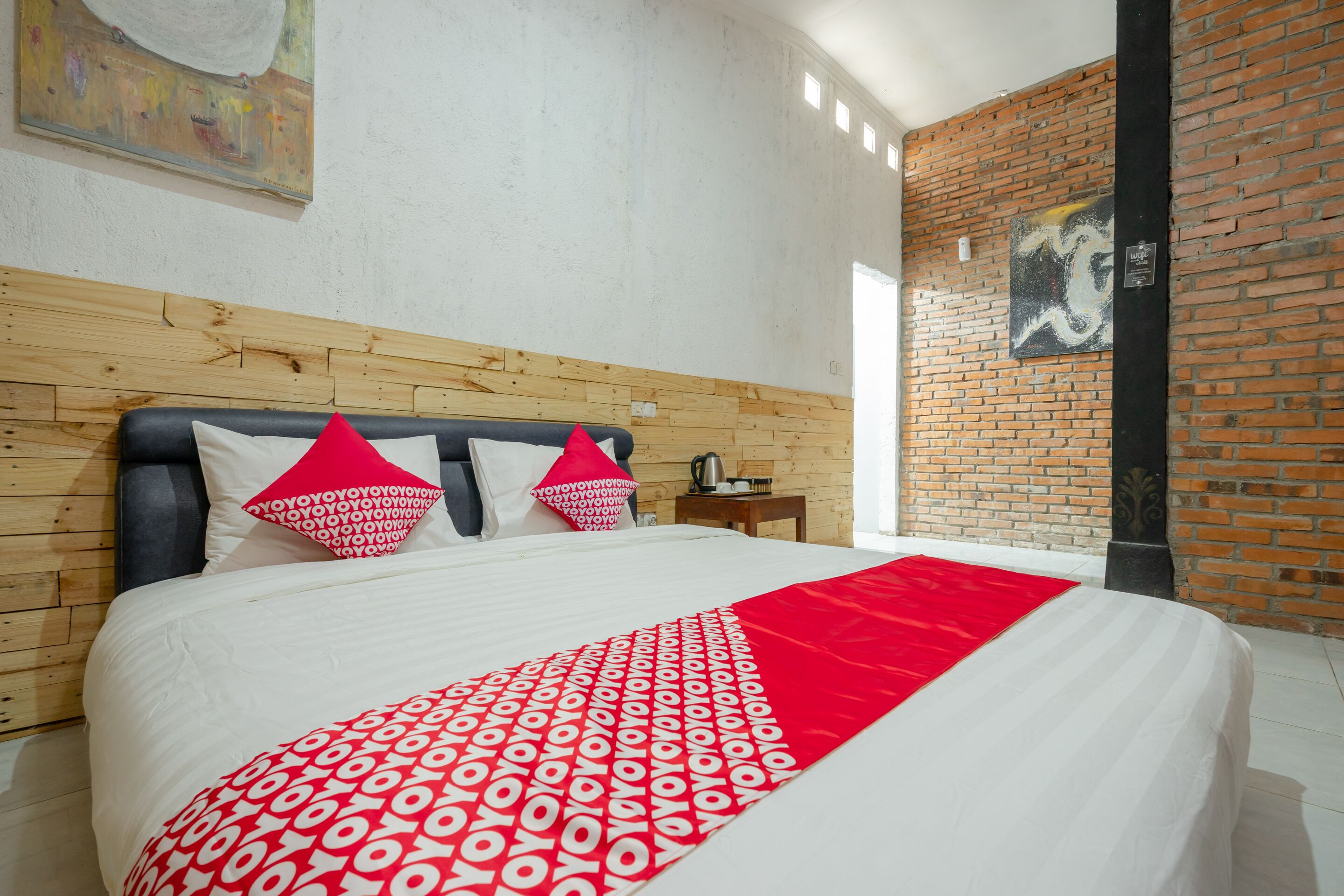 A hotel room with a wooden headboard, a large bed with white bedding and red accents, a red throw blanket, and a brick wall with artwork.