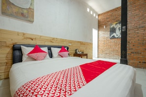 Ein Hotelzimmer mit einem hölzernen Kopfteil, einem großen Bett mit weißer Bettwäsche und roten Akzenten, einer roten Decke und einer Ziegelwand mit Kunstwerken.