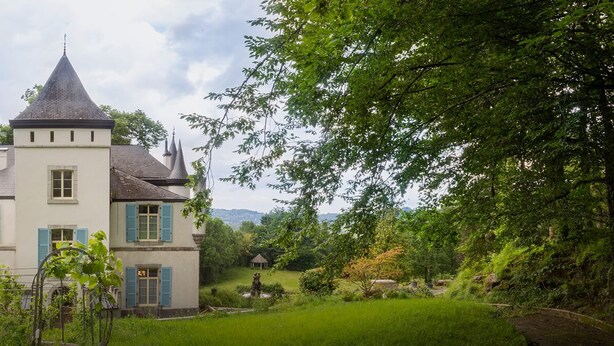 A large, white building with blue shutters and a conical roof, surrounded by lush greenery and trees.