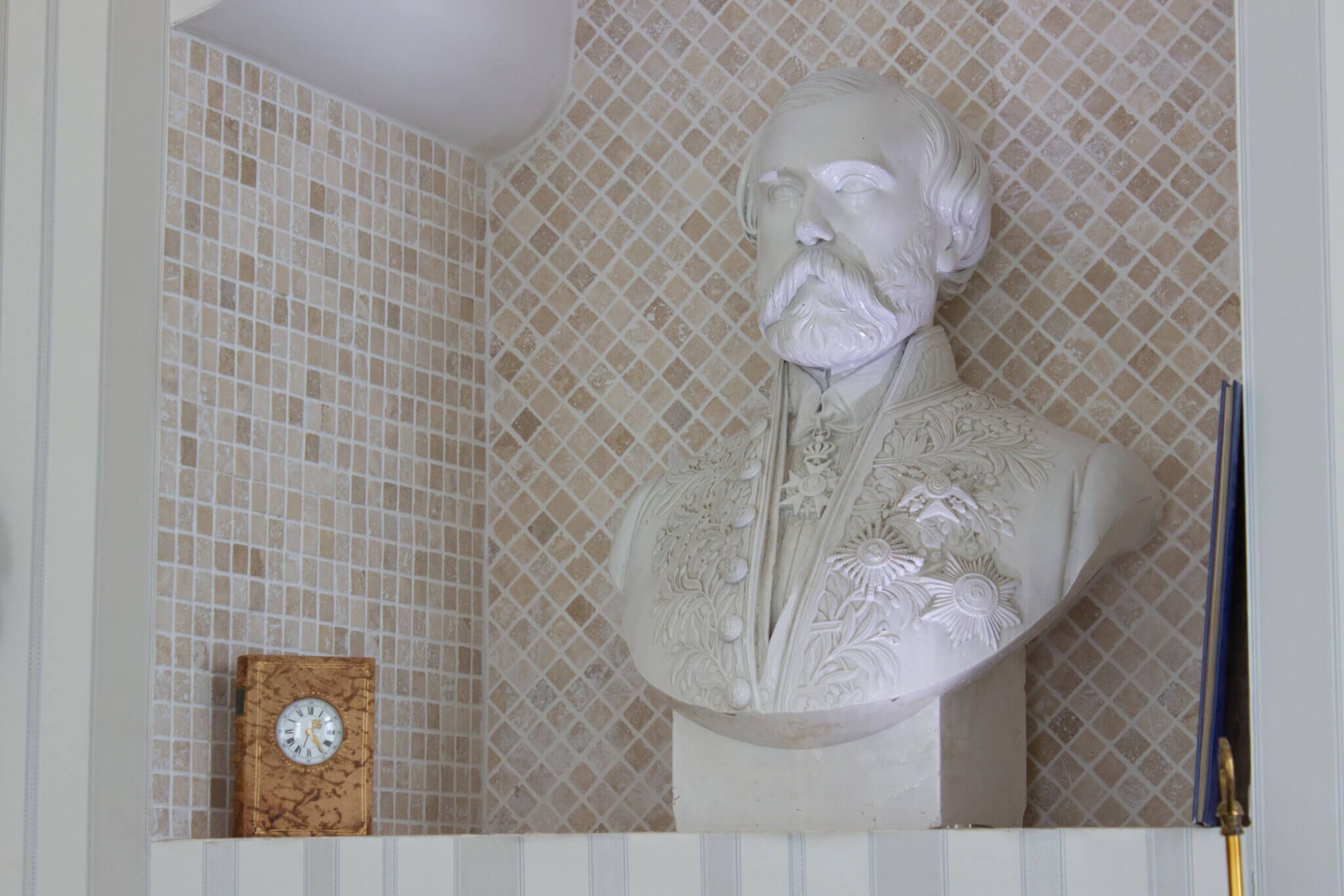 Une sculpture en buste blanc, placée devant un mur recouvert de carreaux, une horloge en bois et un objet bleu.
