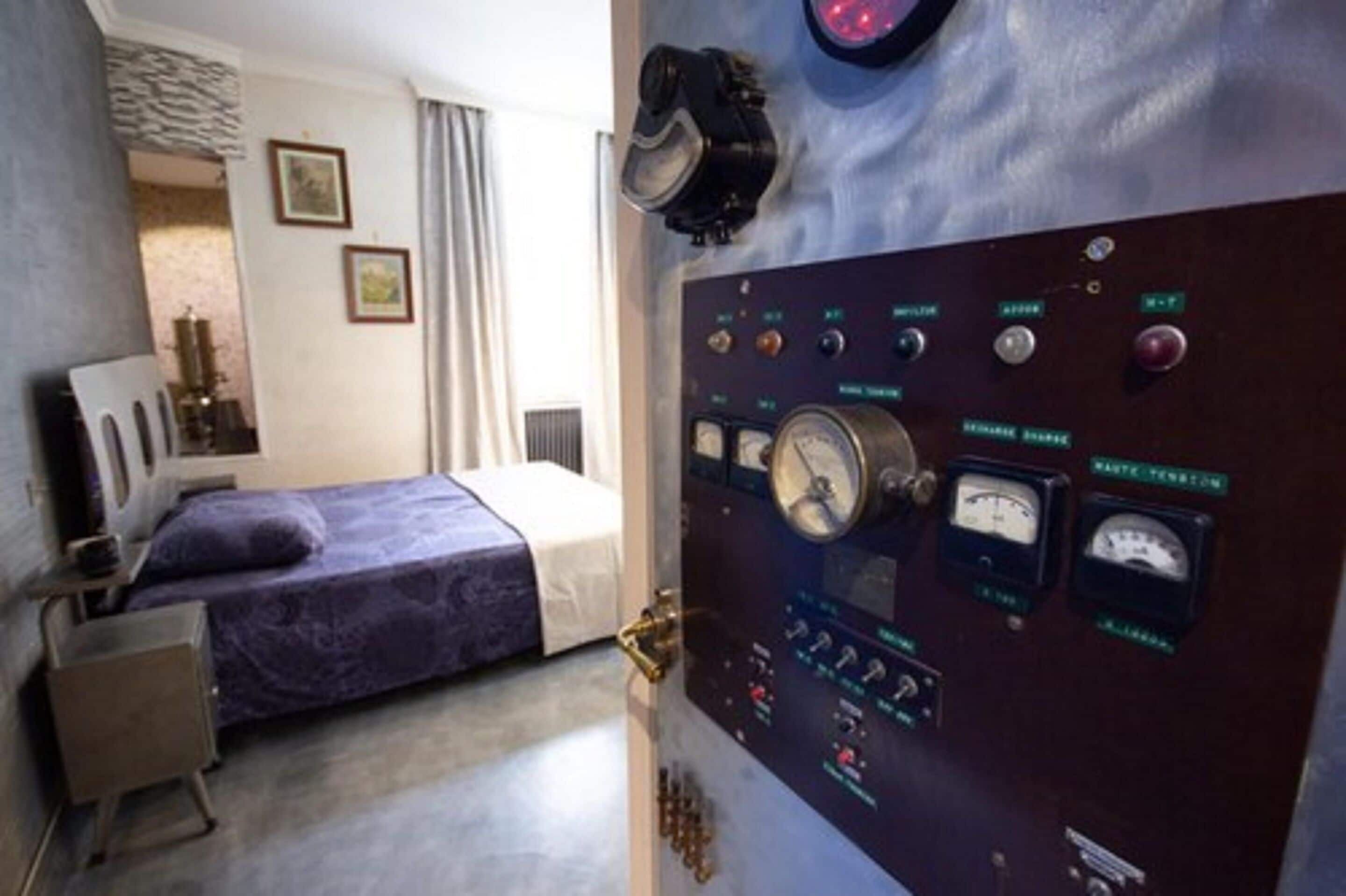 Una habitación con una cama, una mesita pequeña y un panel de control con indicadores y botones.
